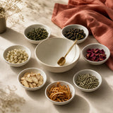 Seven bowls of tea blending ingredients arranged in a circle around an empty mixing bowl and brass scoop
