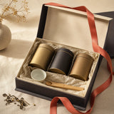 An open premium tea gift box with three tea tins, a porcelain cup and a bamboo scoop, tied with coral ribbon